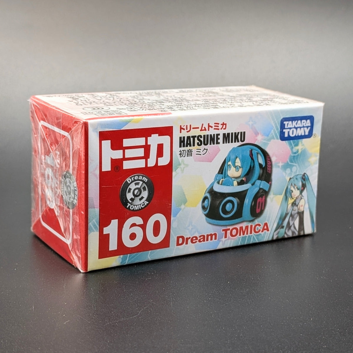 A boxed Dream TOMICA Hatsune Miku toy, shown in its original retail packaging on a dark gray surface. The box features a red and white front panel with the number 160, alongside an illustration of the blue and black character in a car.