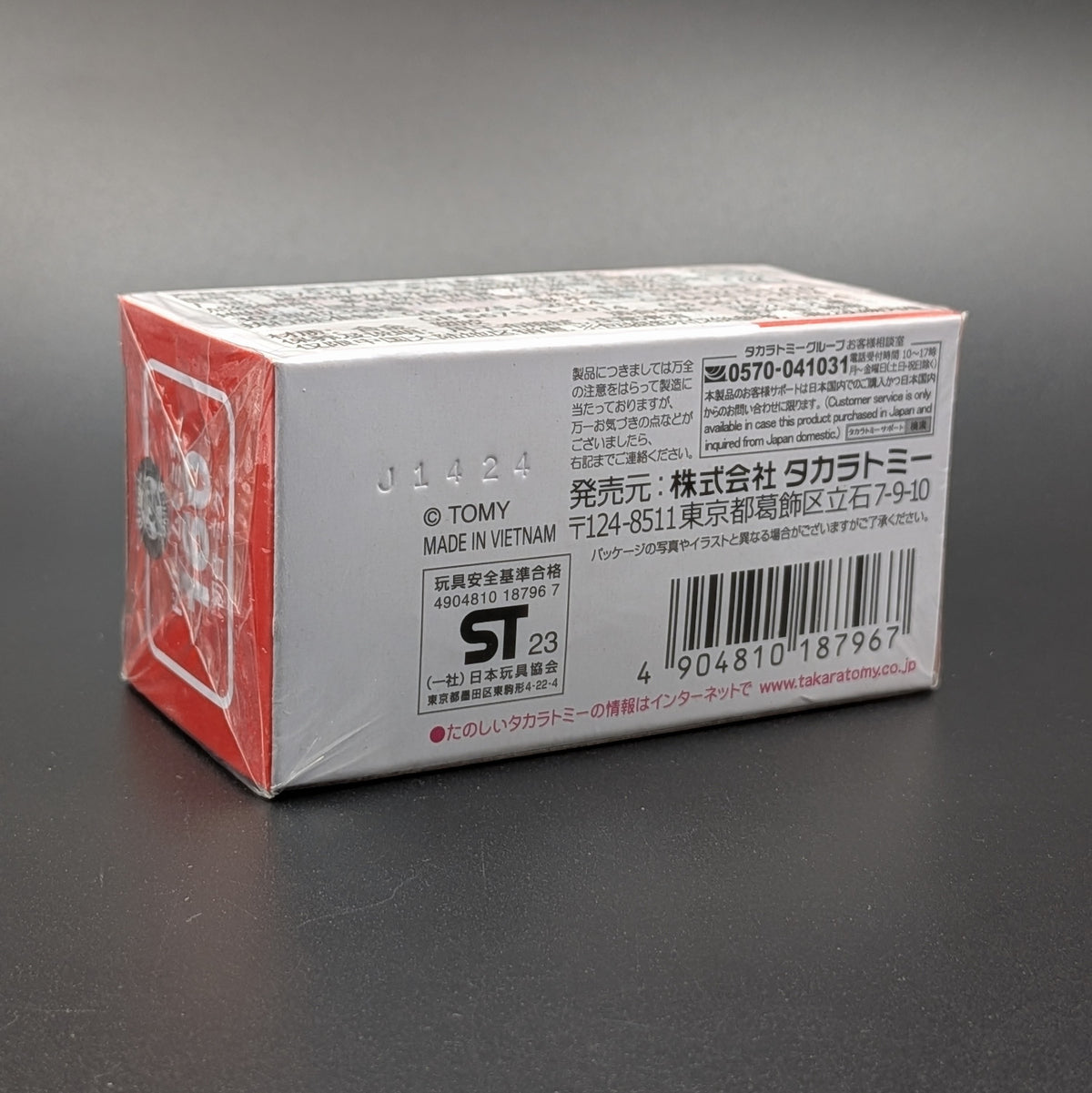 A sealed retail box for a Takara Tomy model, showing the back and side panels with Japanese text, a barcode, and a red graphic on a dark gray background.