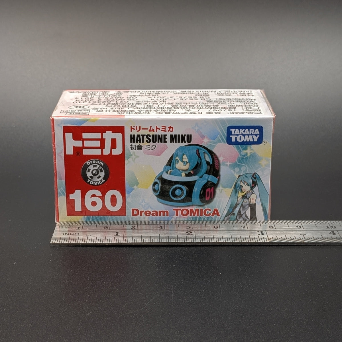 A boxed Tomica Dream Tomica Hatsune Miku model number 160, shown on a dark gray background with a metal ruler underneath for scale.