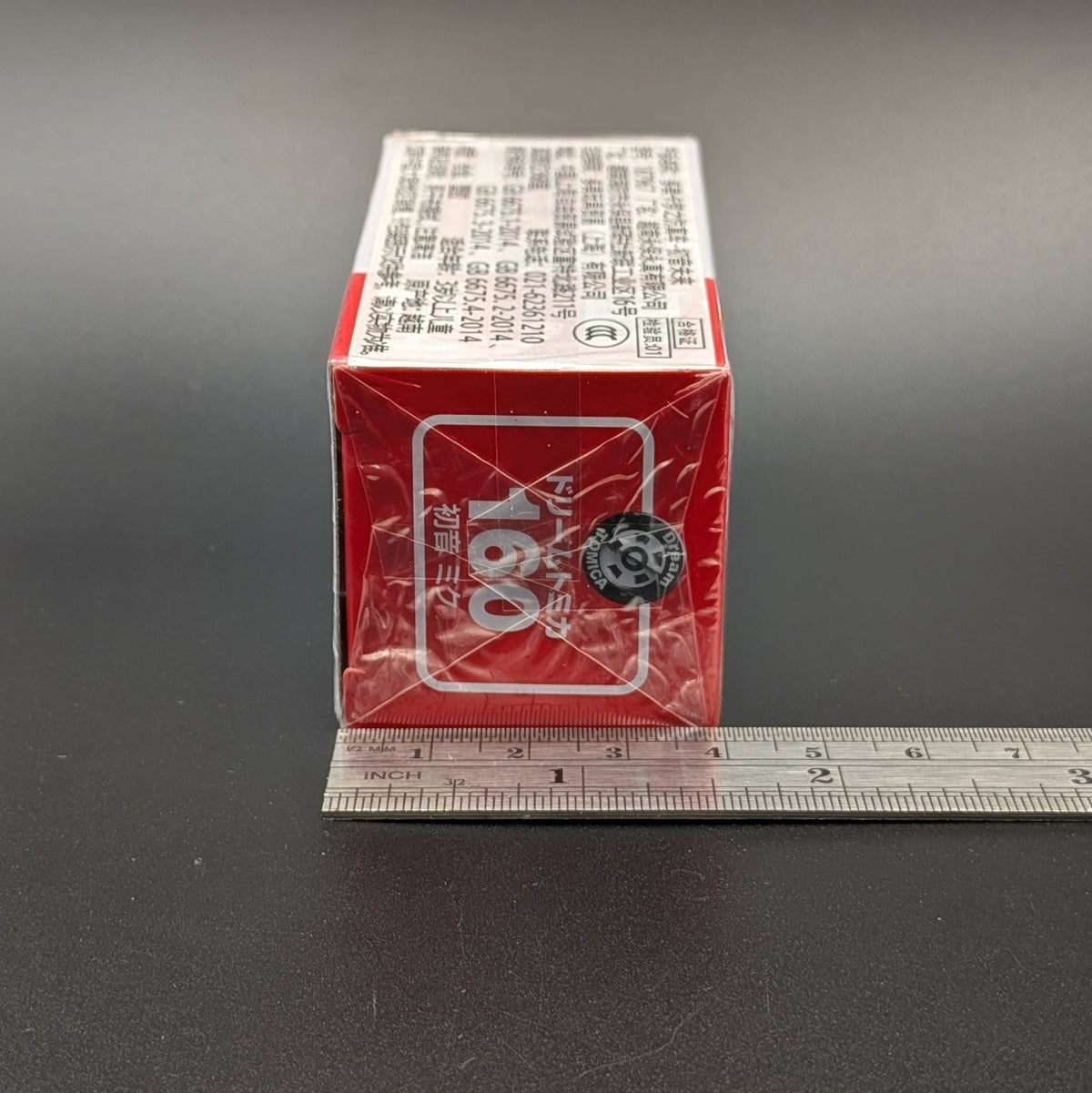 A red and white rectangular product box wrapped in clear plastic, shown on a dark surface with a metal ruler along the bottom edge for scale.