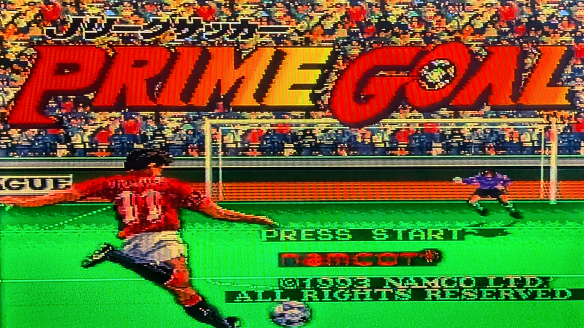 The digital game title screen for Nintendo's J.League Soccer 'Prime Goal' featuring a soccer player kicking a ball toward a goal, with large red title text, a green football field and fans in the stands behind as the background. This shows the SFC cart is tested and working.