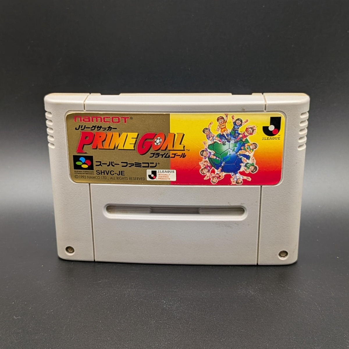 J-League Soccer Prime Goal video game cartridge for Nintendo's Super Famicom (SFC) Entertainment System, shown standing up and facing forwards on a dark grey background. 