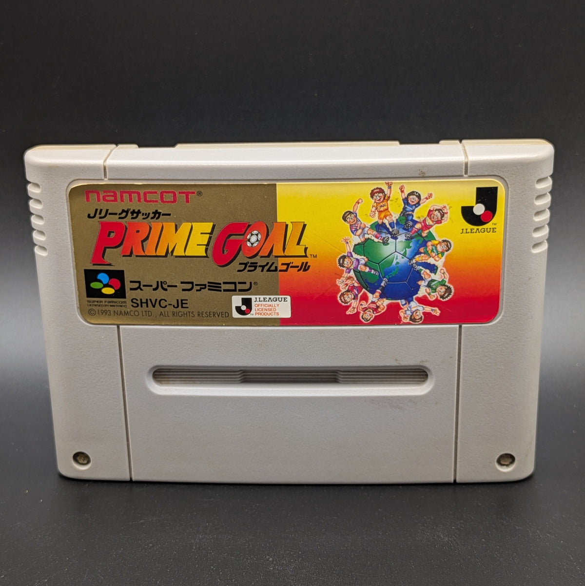 A close up of J-League Soccer Prime Goal video game cartridge for Nintendo's Super Famicom (SFC) Entertainment System, shown standing up and facing forwards on a dark grey background.