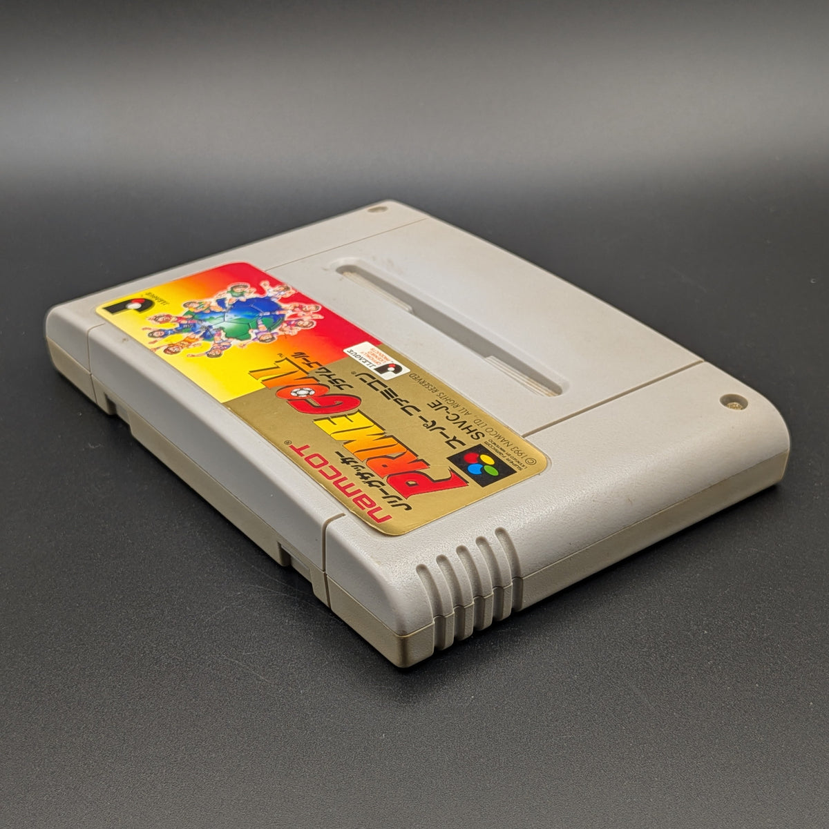 J-League Soccer Prime Goal video game cartridge for Nintendo's Super Famicom (SFC) Entertainment System, shown laying on its back with the label facing upwards. Showing the left hand side and top edge. The cart is displayed on a dark grey background