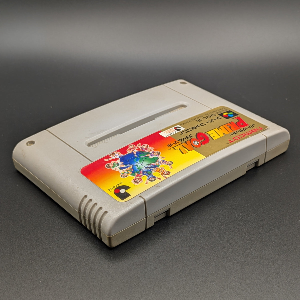 J-League Soccer Prime Goal video game cartridge for Nintendo's Super Famicom (SFC) Entertainment System, shown laying on its back with the label facing upwards. Showing the right hand side and top edge. The cart is displayed on a dark grey background