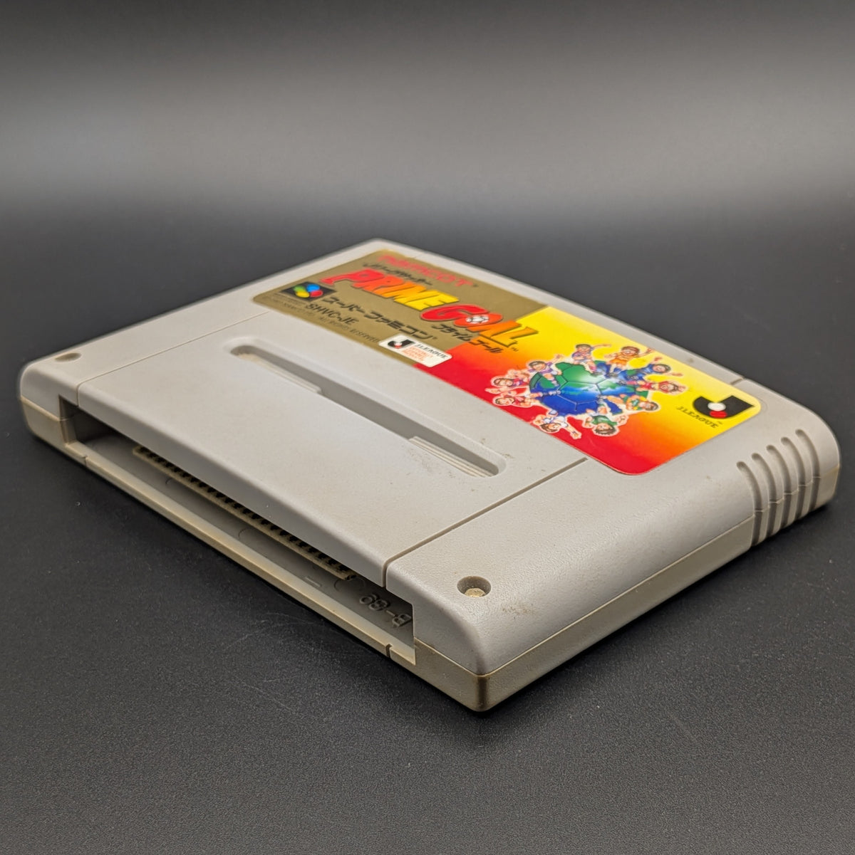 J-League Soccer Prime Goal video game cartridge for Nintendo's Super Famicom (SFC) Entertainment System, shown laying on its back with the label facing upwards. Showing the right hand side and bottom slot. The cart is displayed on a dark grey background