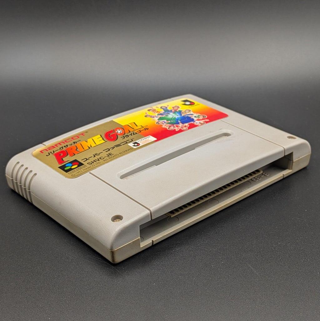 J-League Soccer Prime Goal video game cartridge for Nintendo's Super Famicom (SFC) Entertainment System, shown laying on its back with the label facing upwards. Showing the left hand side and bottom slot. The cart is displayed on a dark grey background