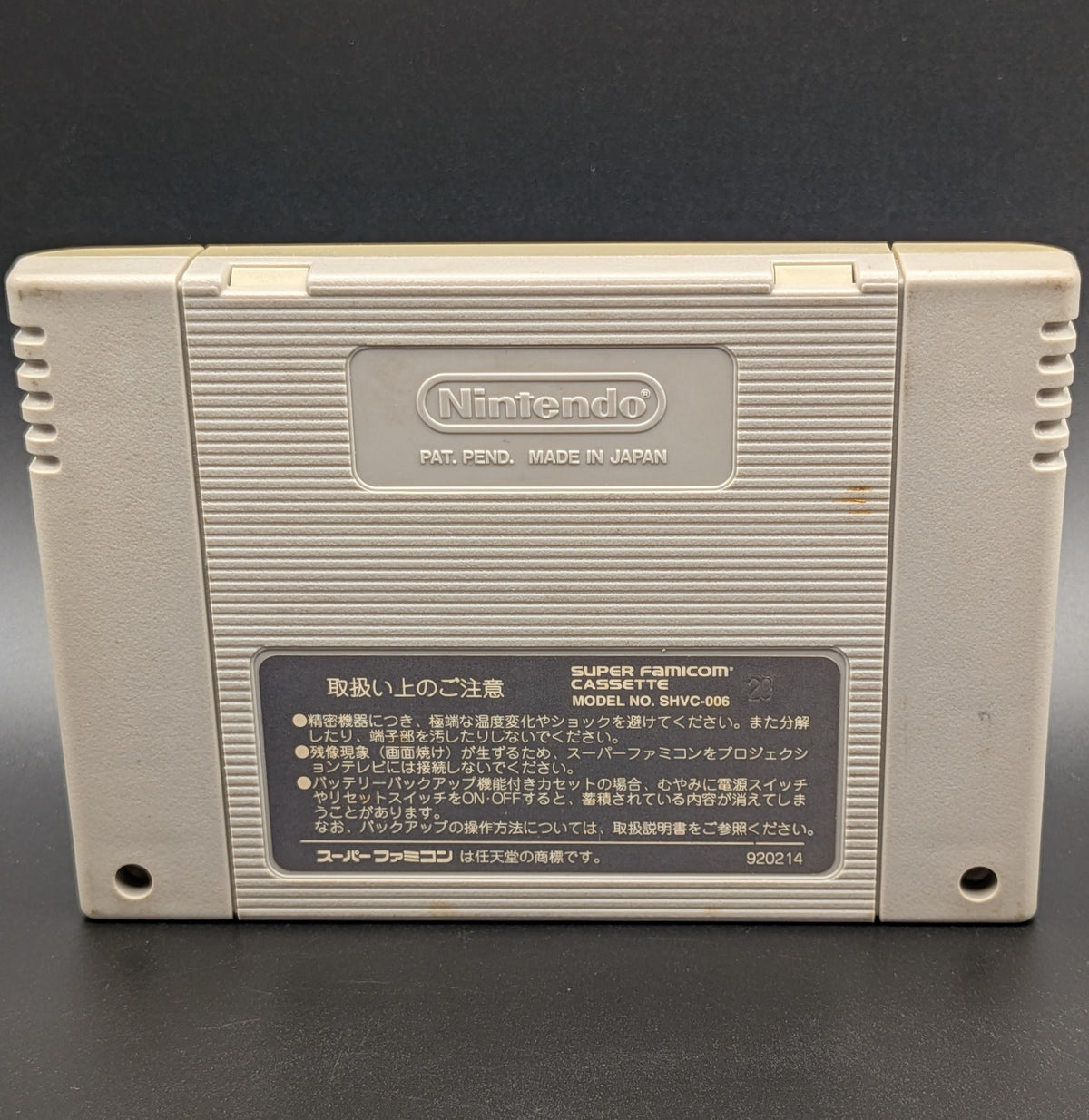 A close up of J League Excite Stage '95 video game cartridge for Nintendo's Super Famicom (SFC) Entertainment System, showing the back of the cart. You can see the back label in Nintendo's stamp. Pictured on a dark grey background. Slight yellowing on the back label.
