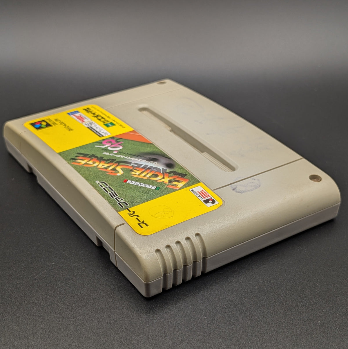 A close up of J League Excite Stage '95 video game cartridge for Nintendo's Super Famicom (SFC) Entertainment System, shown laying on its back with the label facing up and the top edge is visible. Pictured on a dark grey background. Slight yellowing on the front panel.
