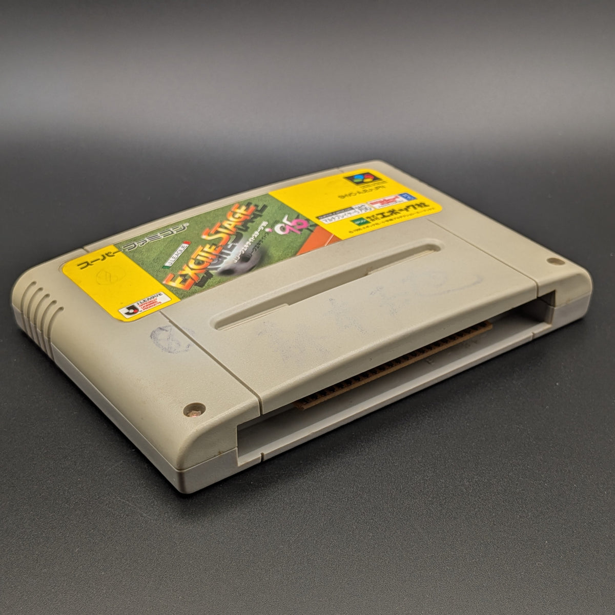 J League Excite Stage '95 video game cartridge for Nintendo's Super Famicom (SFC) Entertainment System, shown laying on its back with the label facing up and the bottom slot is visible. Pictured on a dark grey background.
