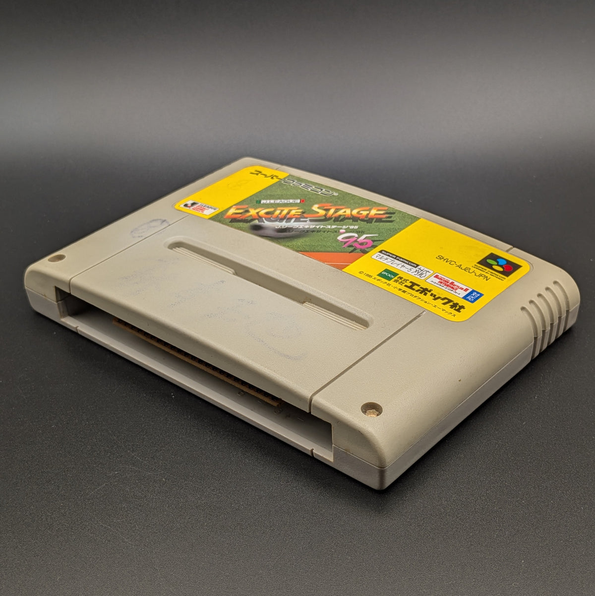 J League Excite Stage '95 video game cartridge for Nintendo's Super Famicom (SFC) Entertainment System, shown laying on its back with the label facing up and the bottom slot is visible. Pictured on a dark grey background.
