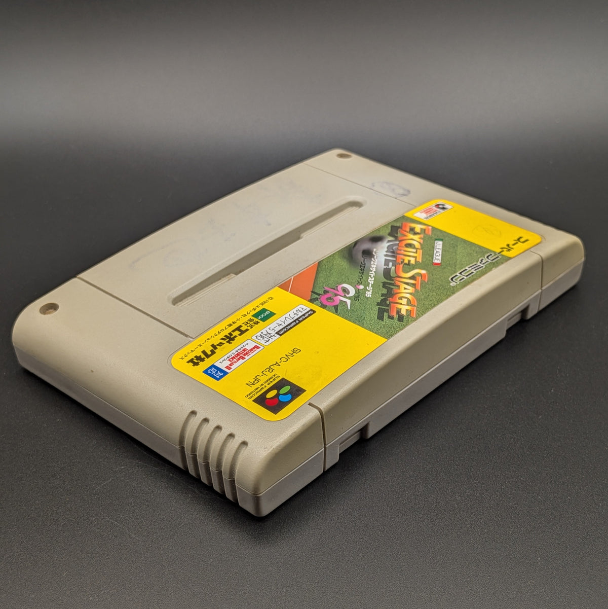 A close up of J League Excite Stage '95 video game cartridge for Nintendo's Super Famicom (SFC) Entertainment System, shown laying on its back with the label facing up and the top edge is visible. Pictured on a dark grey background. Slight yellowing on the front panel.
