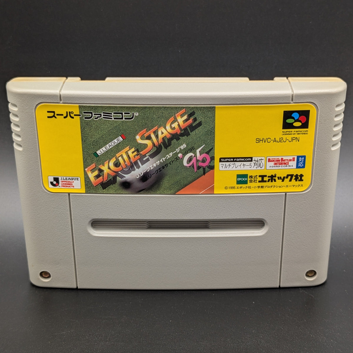 A close up of J-League Excite Stage '95 video game cartridge for Nintendo's Super Famicom (SFC) Entertainment System, shown standing up and facing forward on a dark grey background.