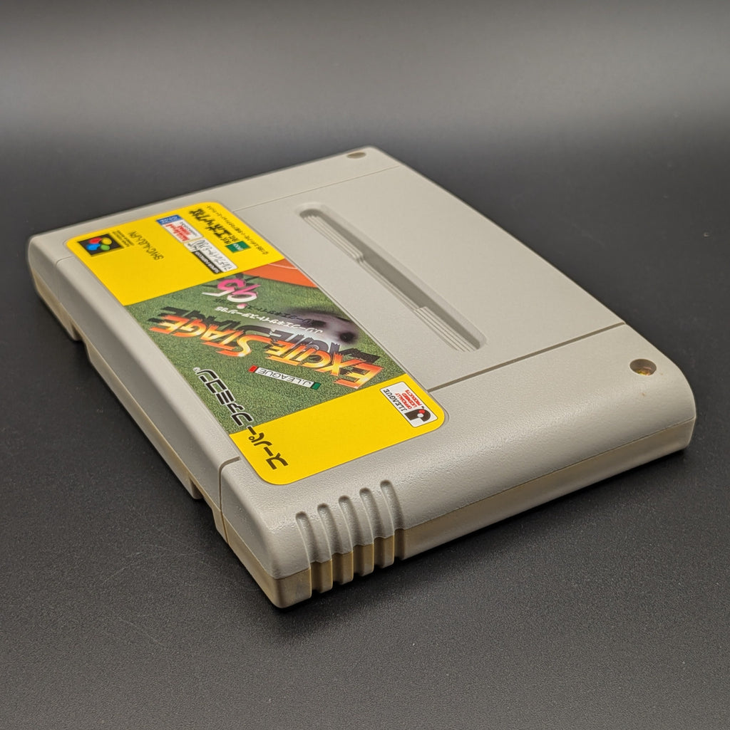 J-League Excite Stage '95 video game cartridge for Nintendo's Super Famicom (SFC) Entertainment System, shown laying on its back with the label facing upwards. Showing the left hand side and top edge. It sits on a dark grey background.