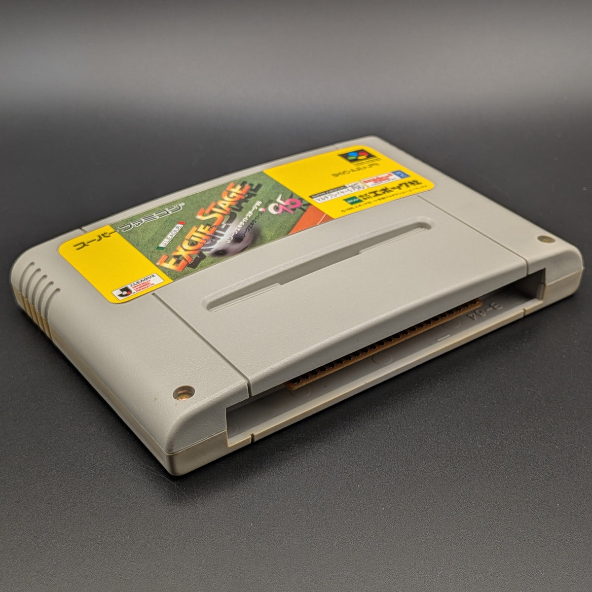 J-League Excite Stage '95 video game cartridge for Nintendo's Super Famicom (SFC) Entertainment System, shown laying on its back with the label facing upwards. Showing the left hand side and bottom slot. It sits on a dark grey background.