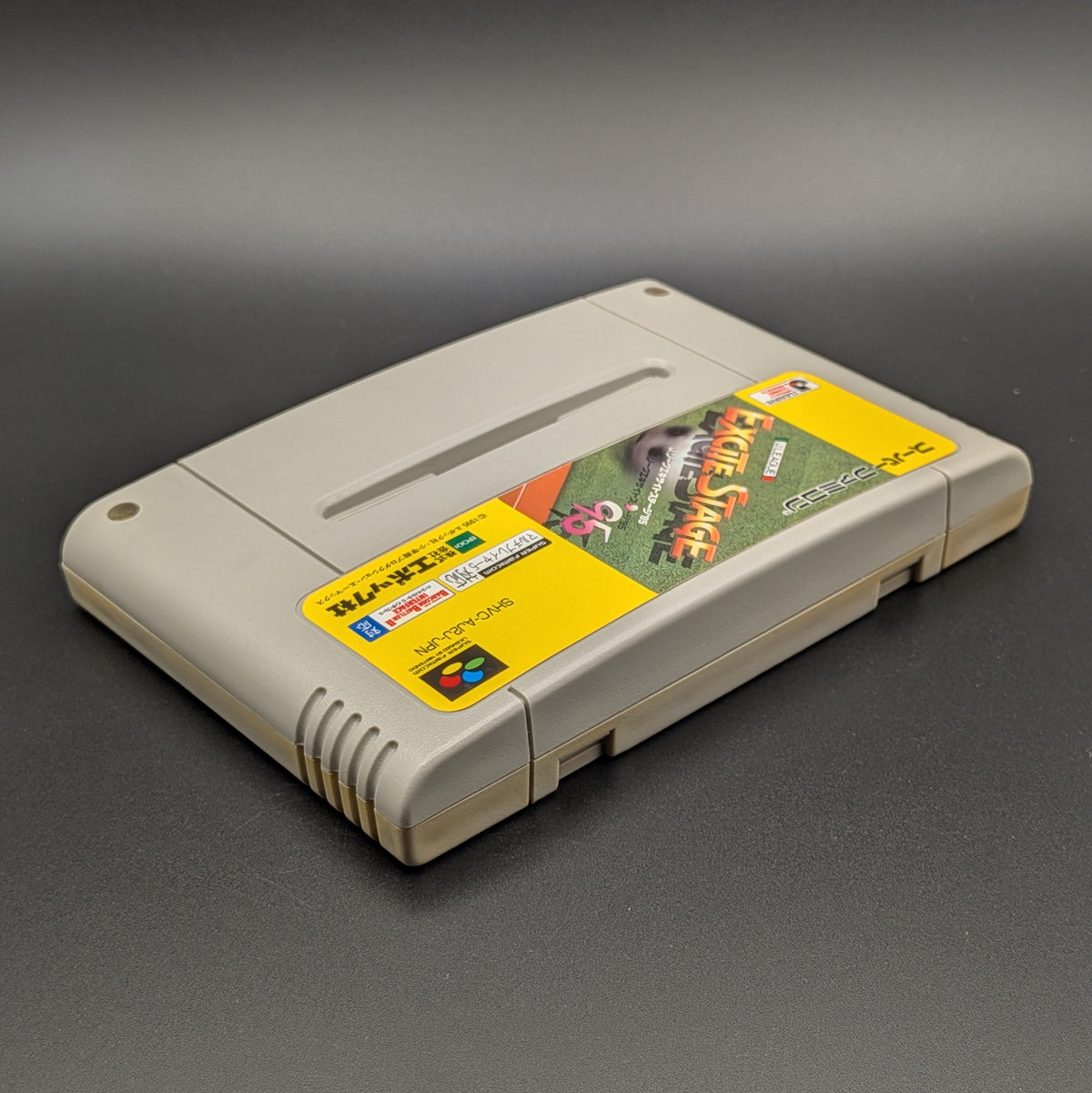 J-League Excite Stage '95 video game cartridge for Nintendo's Super Famicom (SFC) Entertainment System, shown laying on its back with the label facing upwards. Showing the right hand side and top edge. It sits on a dark grey background.