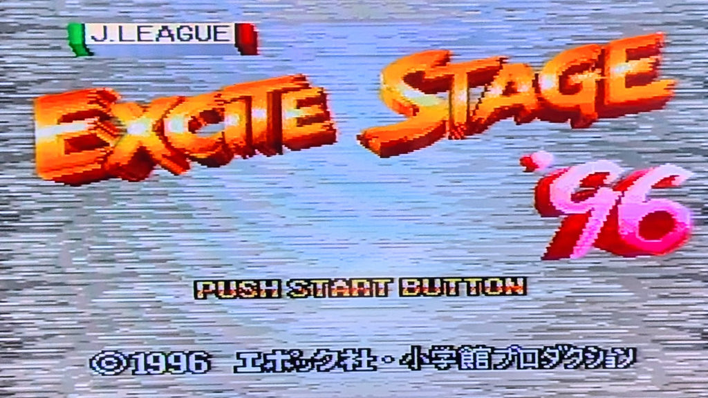 Digital title screen for the video game 'Excite Stage '96' showing stylized orange 3D text, a pink '96, and 'J.LEAGUE' at the top left hand corner, on a blue-gray textured background.