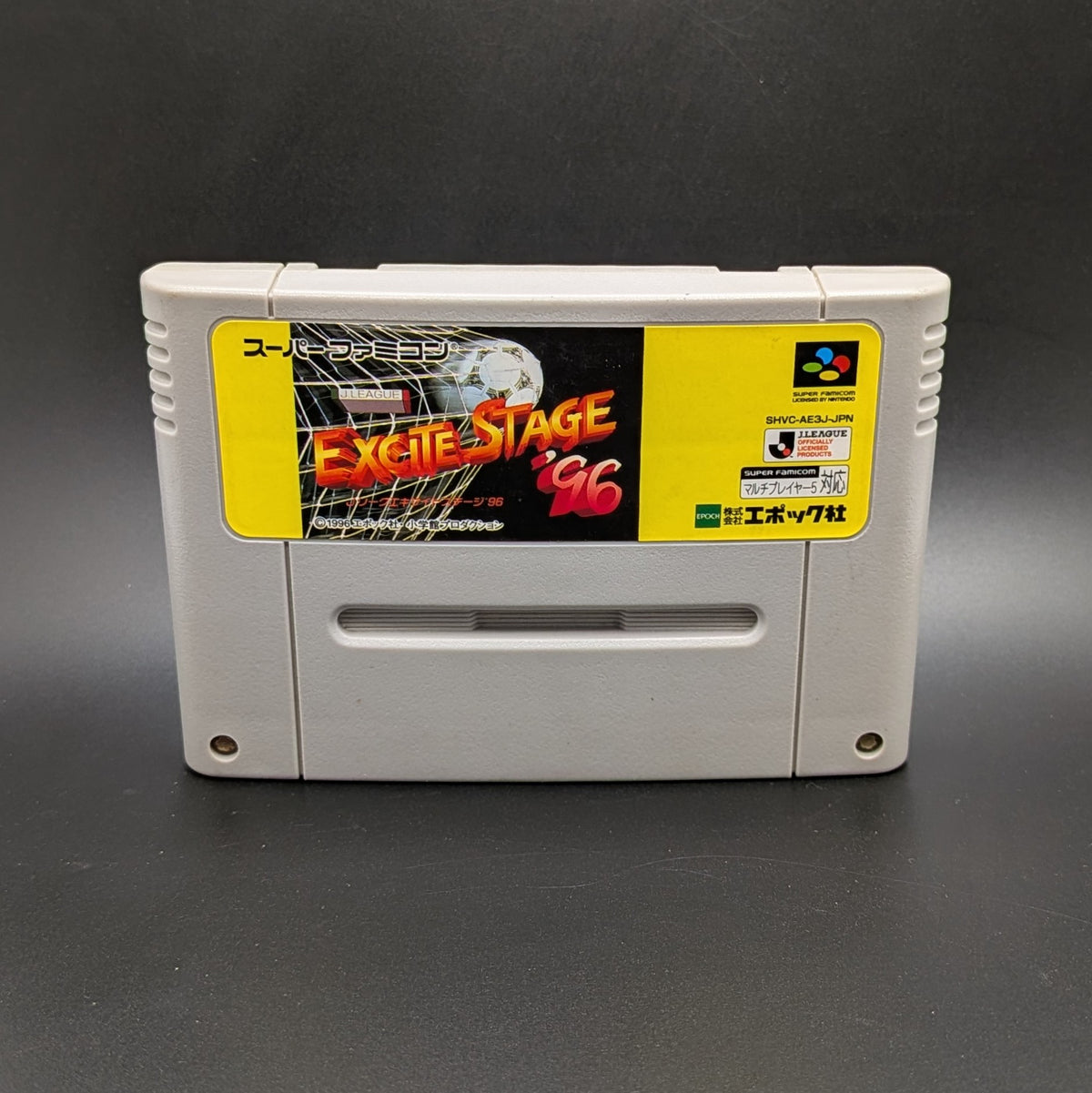 J.League Excite Stage '96 video game cartridge for the Super Famicom Entertainment System, shown standing up and facing forward on a dark gray background.
