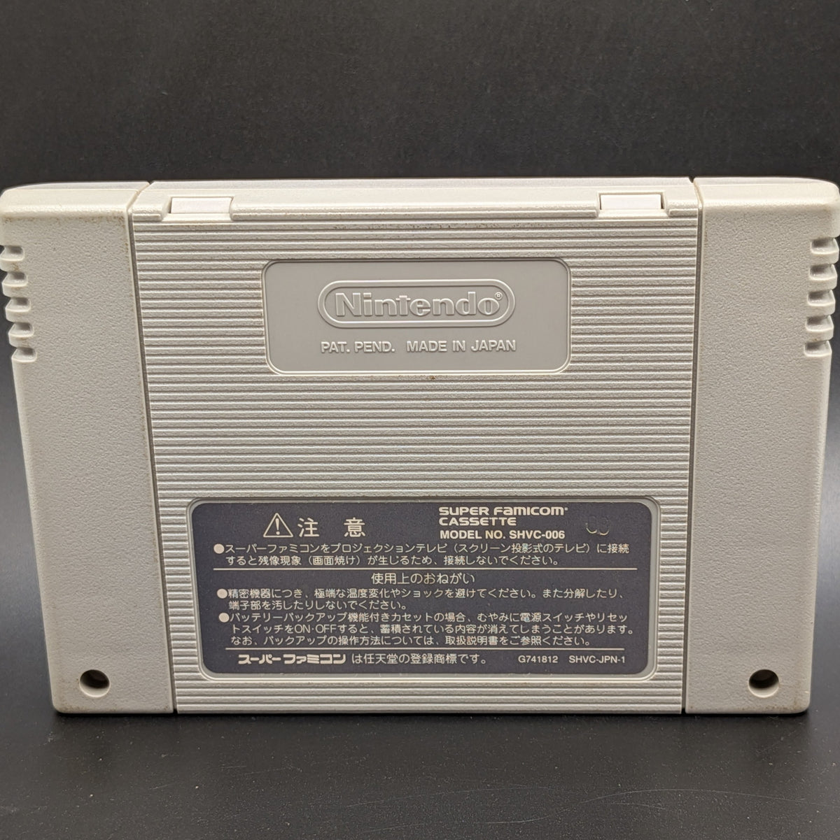 The back of the video game cartridge for  J.League Excite Stage '96 for the Super Famicom. It shows the imprinted Nintendo logo and back label. Pictured on a dark grey background.