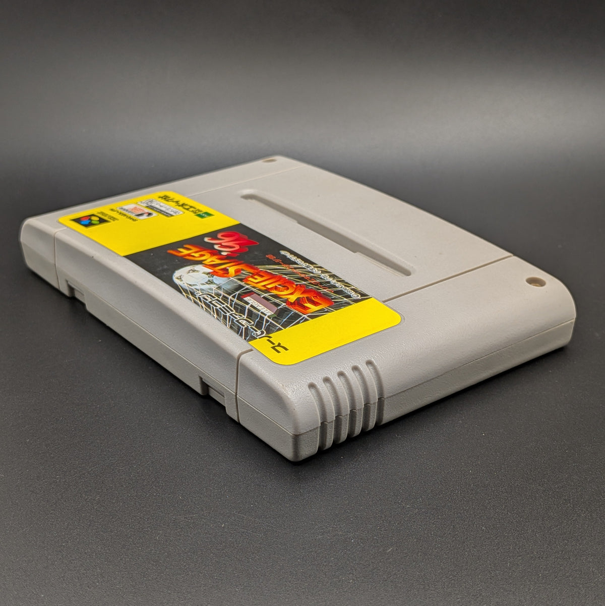 J.League Excite Stage '96 video game cartridge for the Super Famicom Entertainment System, shown laying on its back, at a 3/4 angle facing up so the label is visible. Pictured on a dark grey background.