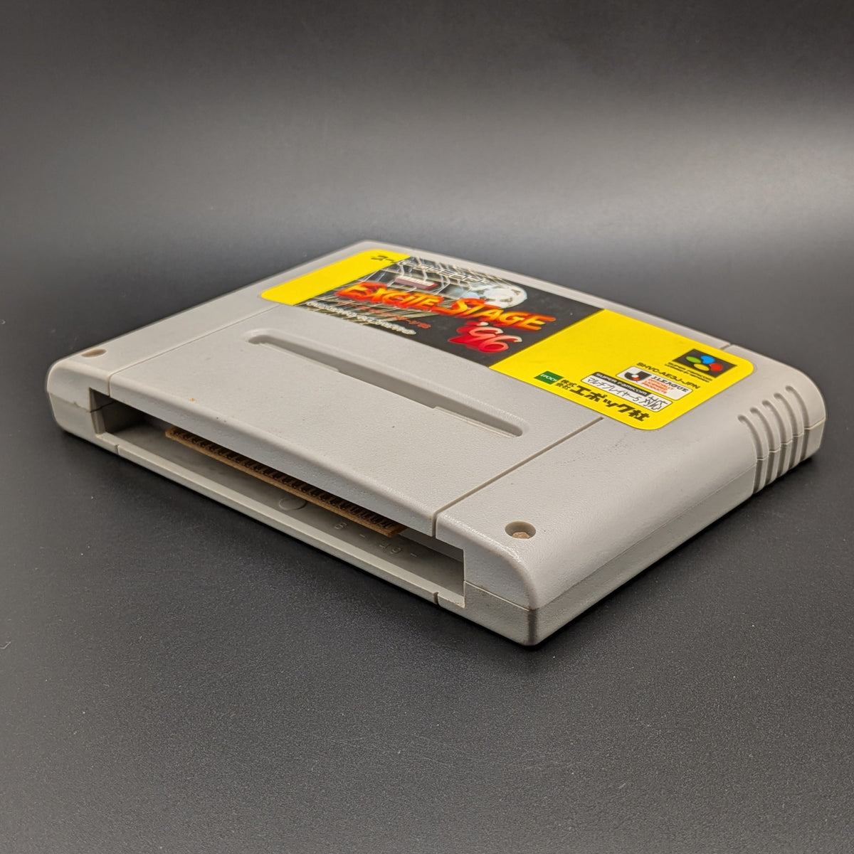 J.League Excite Stage '96 video game cartridge for the Super Famicom Entertainment System, shown laying on its back with the label facing up, at a 3/4 angle. Pictured on a dark grey background.
