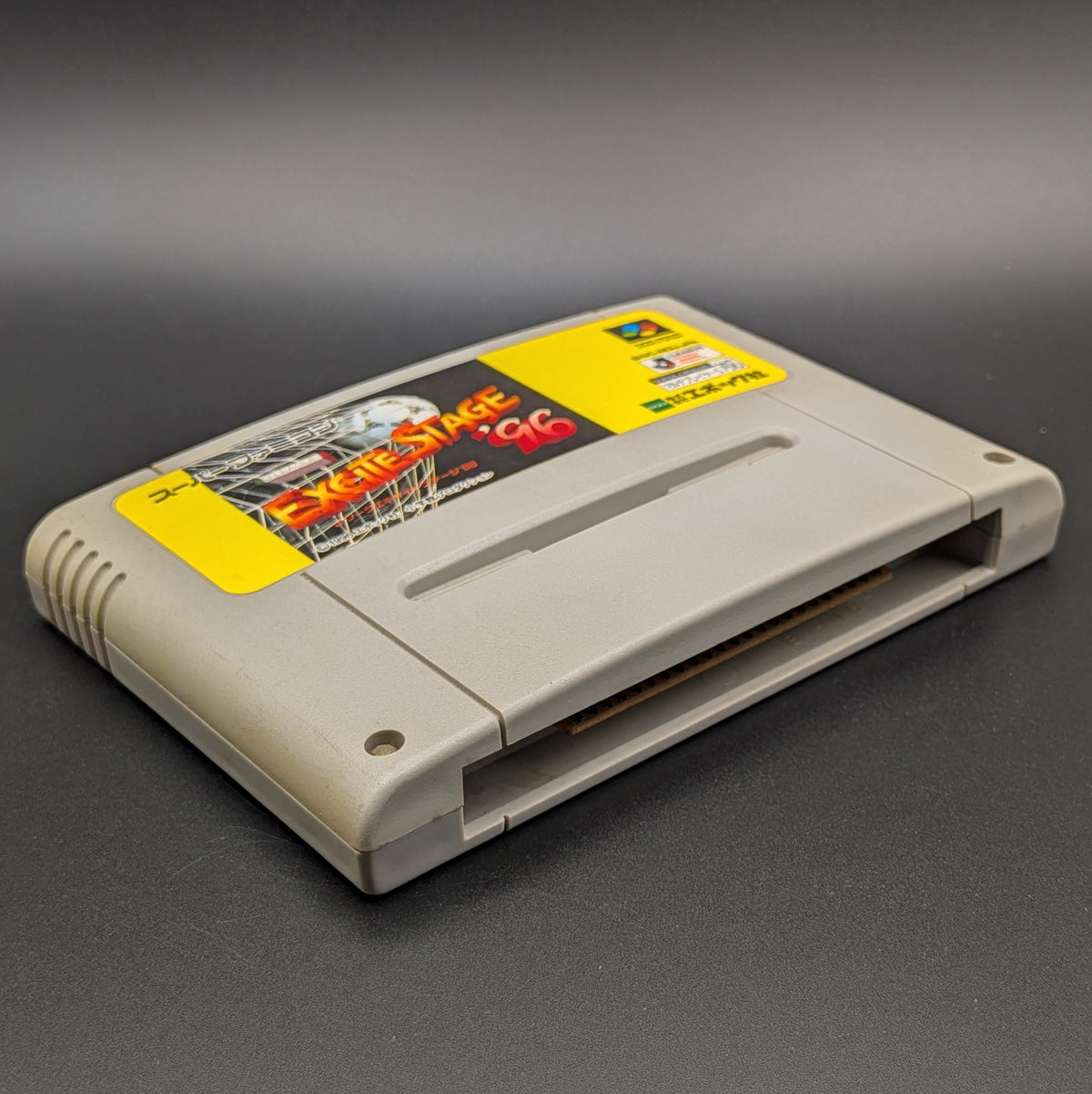 J League Excite Stage '96 video game cartridge for Nintendo's Super Famicom (SFC) Entertainment System, shown laying on its back with the label facing upwards. Showing the left hand side and bottom slot. The cart is displayed on a dark grey background