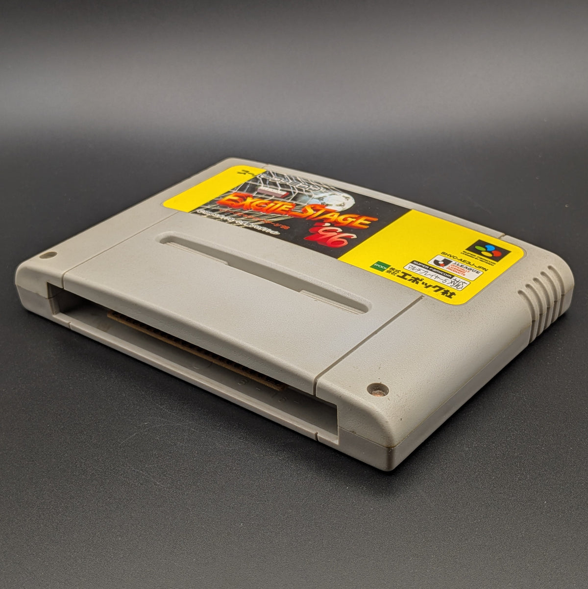 J League Excite Stage '96 video game cartridge for Nintendo's Super Famicom (SFC) Entertainment System, shown laying on its back with the label facing upwards. Showing the right hand side and bottom slot. The cart is displayed on a dark grey background