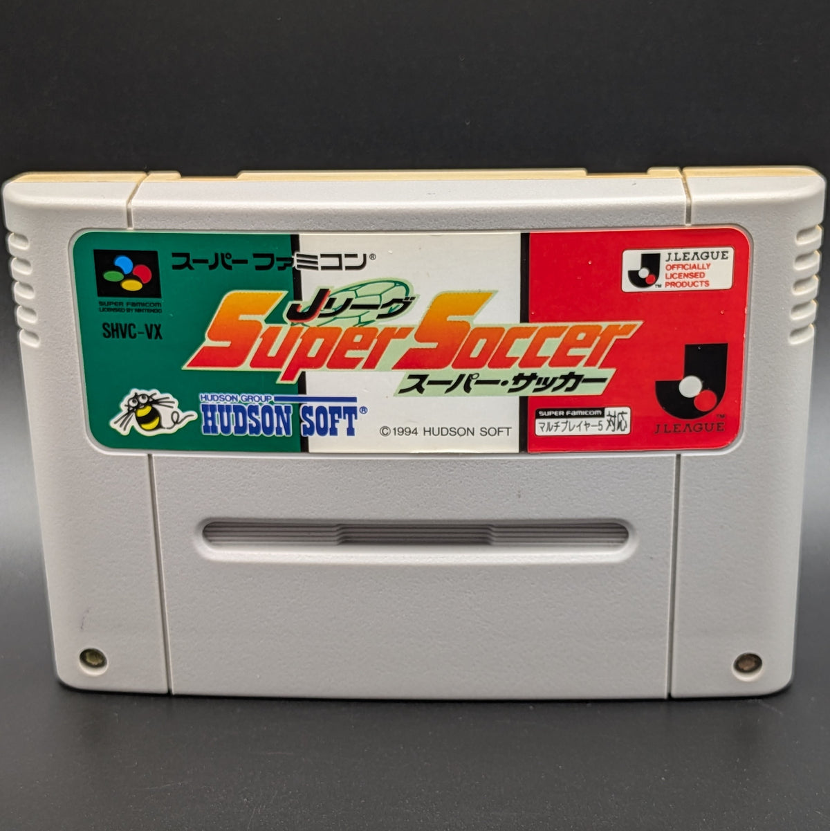 A close up of J League Super Soccer video game cartridge for Nintendo's Super Famicom (SFC) Entertainment System, shown standing up and facing forwards on a dark grey background.