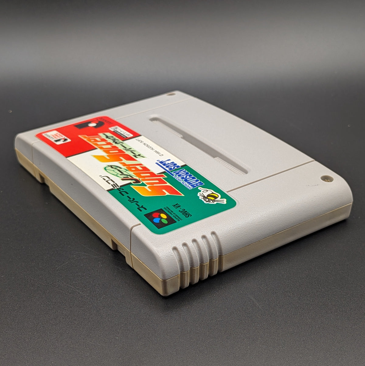 J League Super Soccer video game cartridge for Nintendo's Super Famicom (SFC) Entertainment System, shown laying on its back with the label facing upwards. Showing the left hand side and top slot. It sits on a dark grey background.