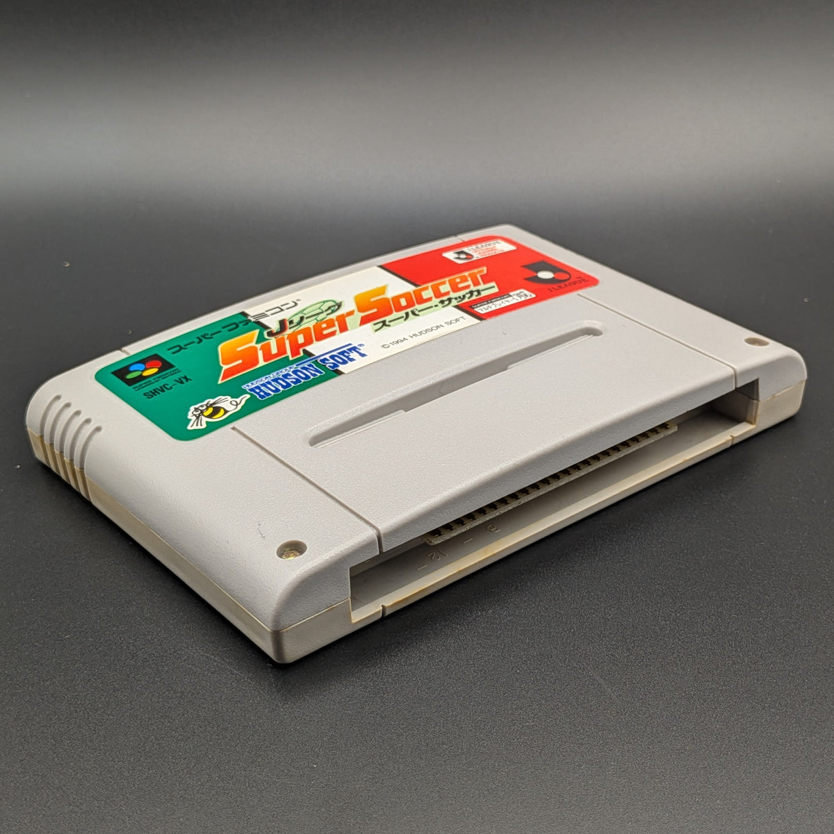 J League Super Soccer video game cartridge for Nintendo's Super Famicom (SFC) Entertainment System, shown laying on its back with the label facing upwards. Showing the left hand side and bottom slot. It sits on a dark grey background.