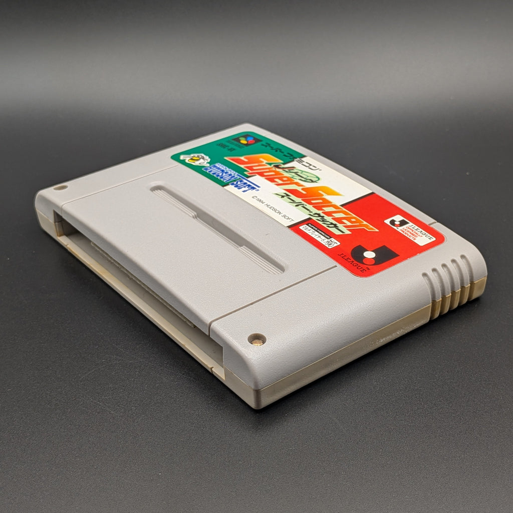 J League Super Soccer video game cartridge for Nintendo's Super Famicom (SFC) Entertainment System, shown laying on its back with the label facing upwards. Showing the right hand side and bottom slot. It sits on a dark grey background.