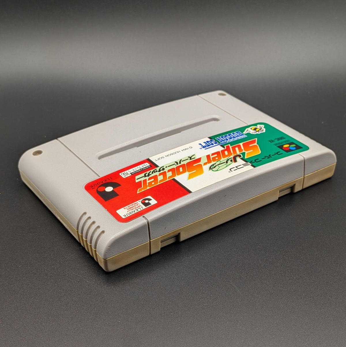J League Super Soccer video game cartridge for Nintendo's Super Famicom (SFC) Entertainment System, shown laying on its back with the label facing upwards. Showing the right hand side and top slot. It sits on a dark grey background.