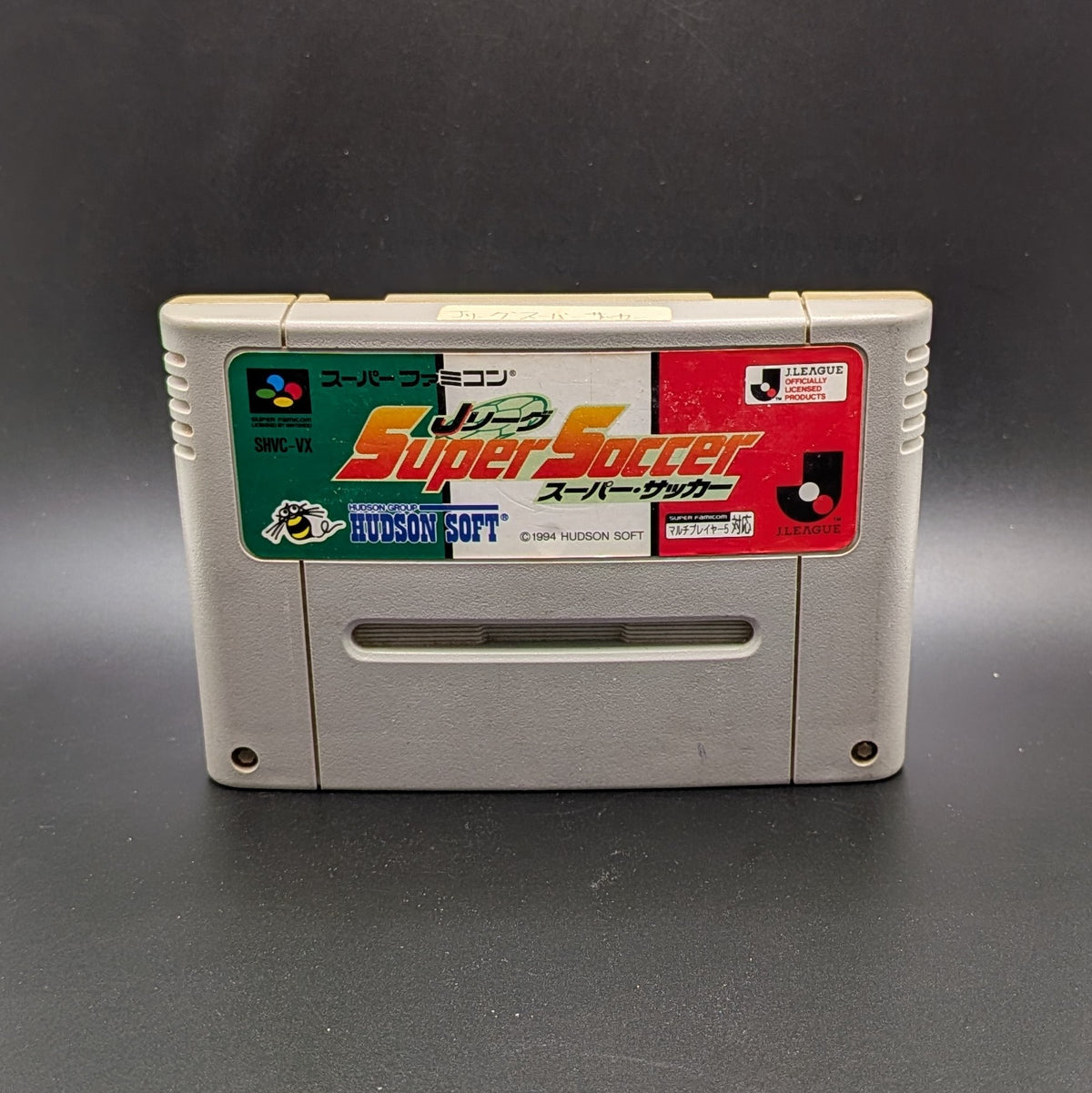 J League Super Soccer video game cartridge for Nintendo's Super Famicom (SFC) Entertainment System, shown standing up and facing forward on a dark grey background.
