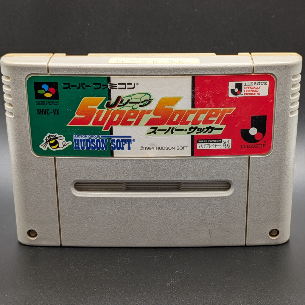 A close up of J League Super Soccer video game cartridge for Nintendo's Super Famicom (SFC) Entertainment System, shown standing up and facing forward on a dark grey background.
