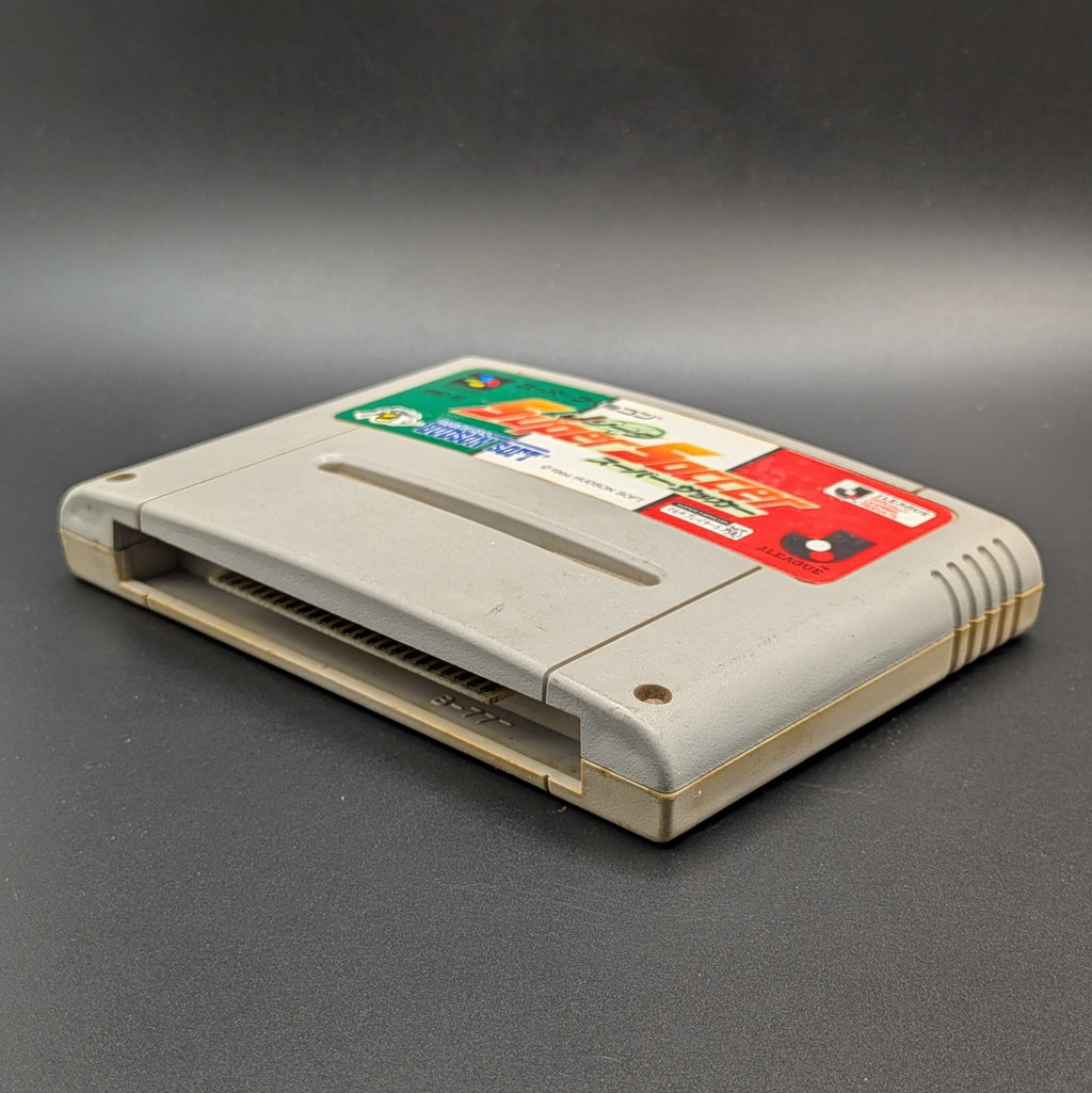 J League Super Soccer video game cartridge for Nintendo's Super Famicom (SFC) Entertainment System, shown laying on its back with the green, white, and red label facing up on a dark grey background.
