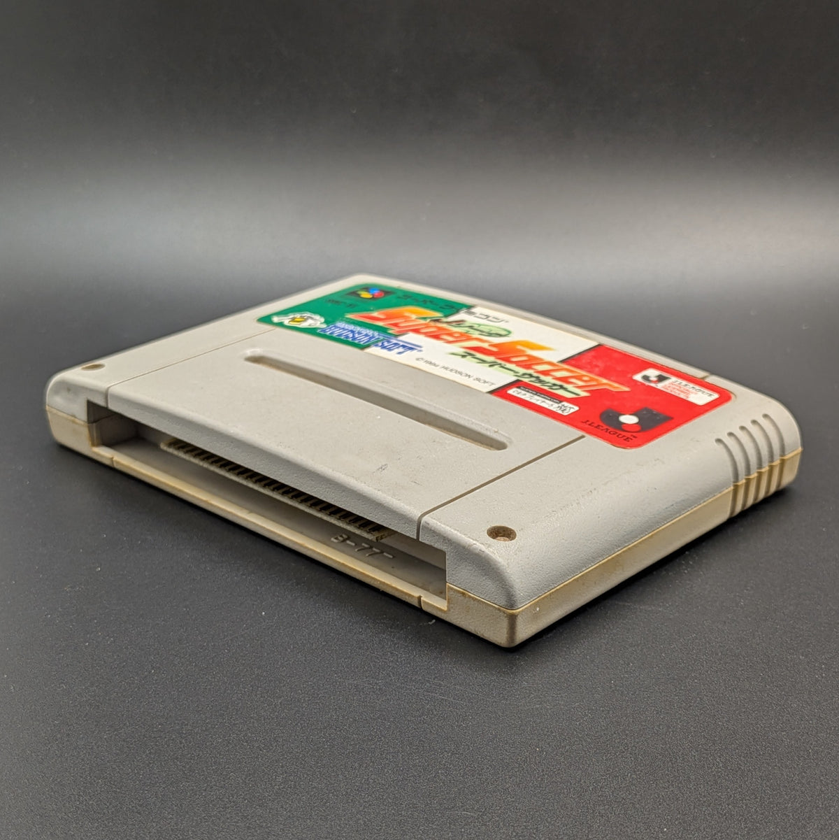 J League Super Soccer video game cartridge for Nintendo's Super Famicom (SFC) Entertainment System, shown laying on its back with the green, white, and red label facing up on a dark grey background.
