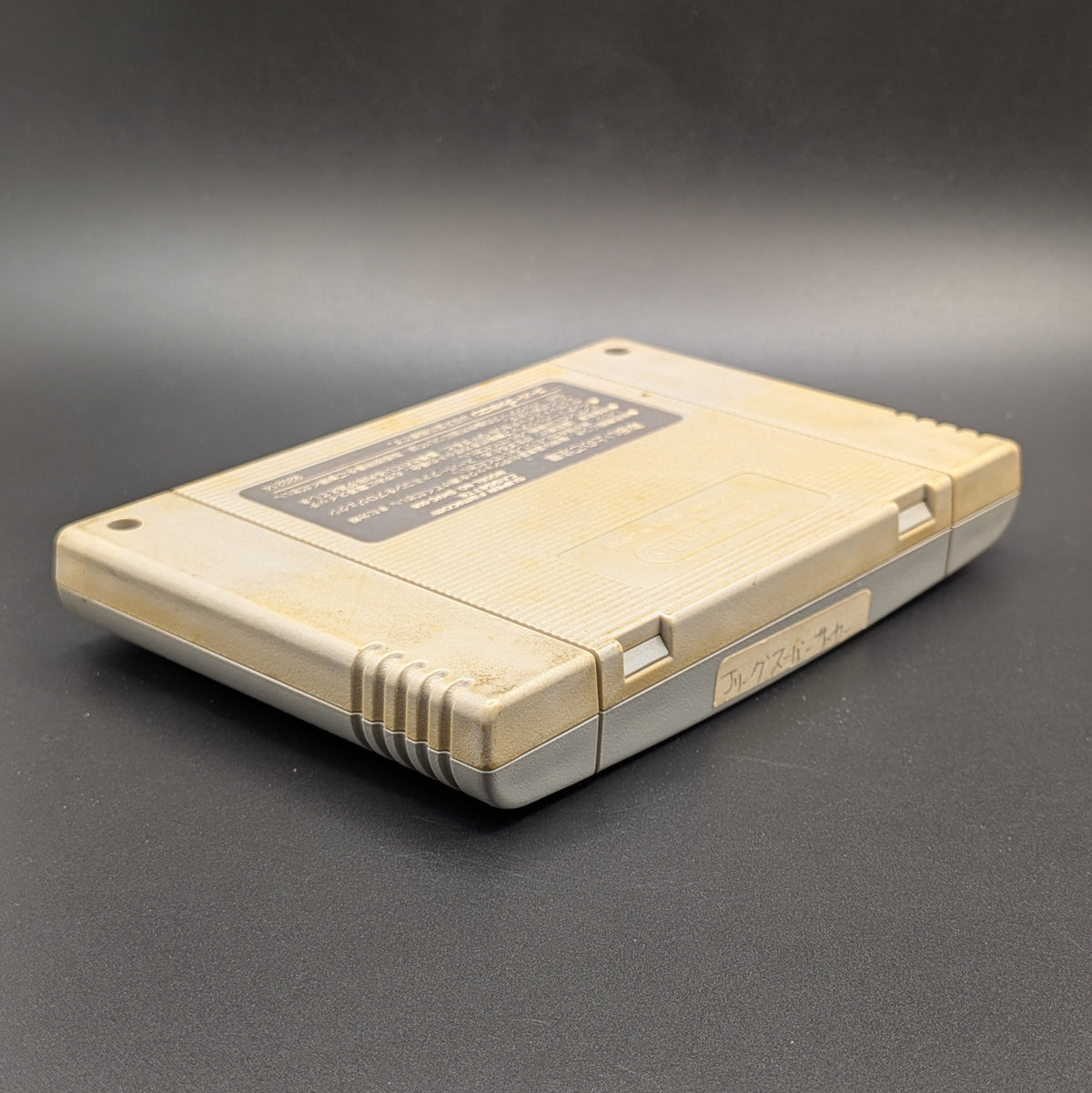 J League Super Soccer video game cartridge for Nintendo's Super Famicom (SFC) Entertainment System, shown from an angled top-down view on a dark grey background. You can see yellowing on the back panel and a hand-written Japanese sticker on the top edge.