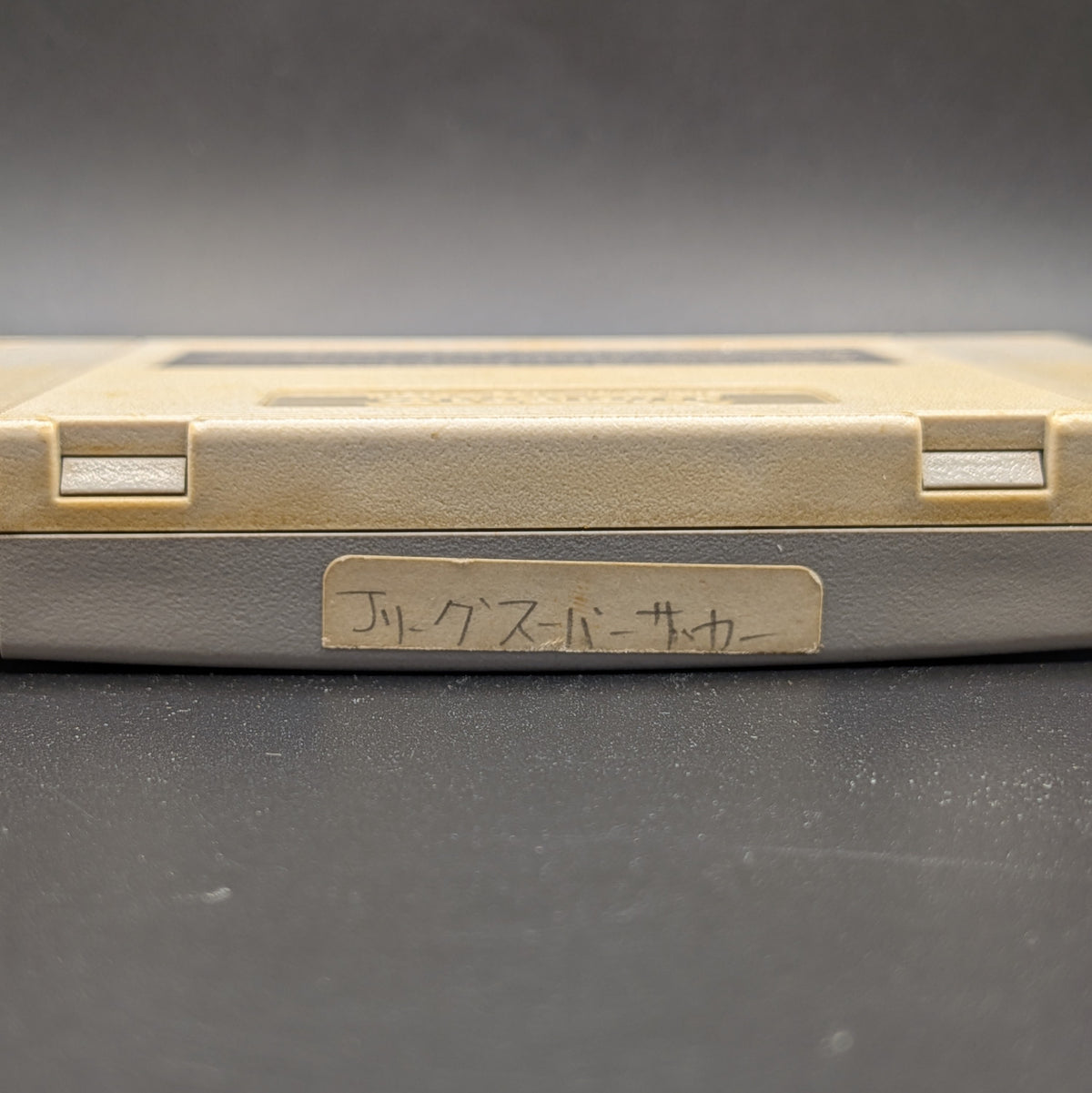 J League Super Soccer video game cartridge for Nintendo's Super Famicom (SFC) Entertainment System, showing a close up of the hand-written Japanese sticker on the top edge.