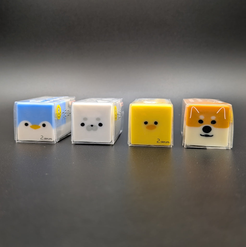 Four small square character-shaped eraser in clear plastic packaging, lined up on a dark gray surface against a dark gray background. From left to right, the faces are a blue penguin, a white seal, a yellow duck, and an orange Shiba Inu dog.