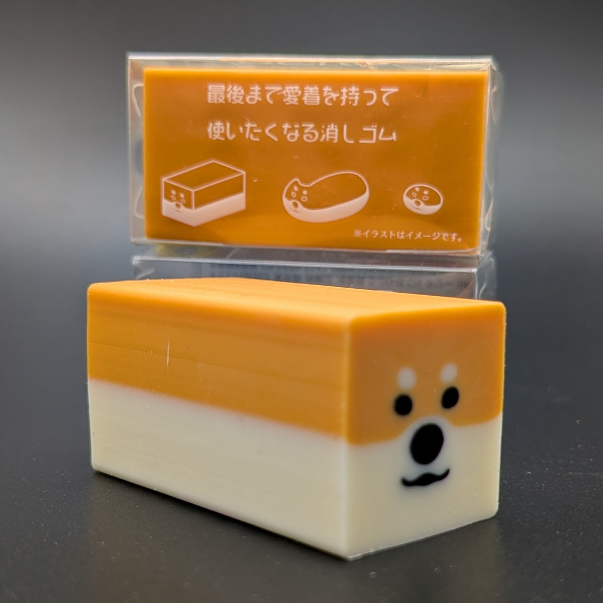 A small orange and white rectangular character-shaped eraser with a cute shiba inu face on the end. The side of the package can be seen in the background, showing an image guide of how the eraser works and changes shape over time.