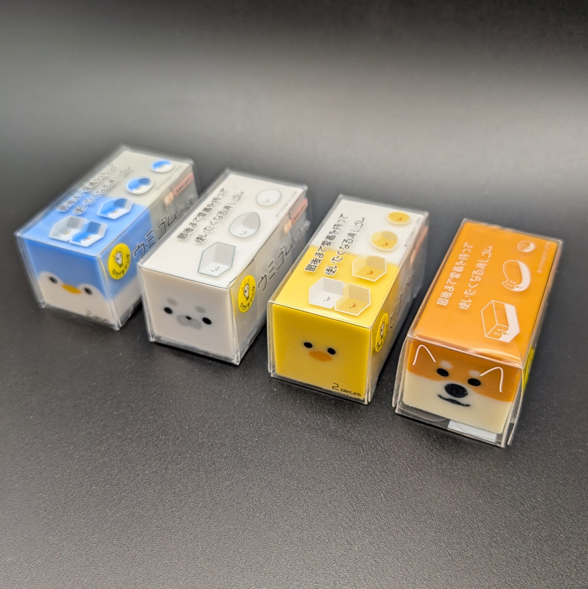 Four small square character-shaped erasers in clear plastic packaging, with Japanese text on the tops. Arranged in a row on a dark grey surface, from left to right, the faces are a blue penguin, a white seal, a yellow duck, and an orange Shiba Inu dog.