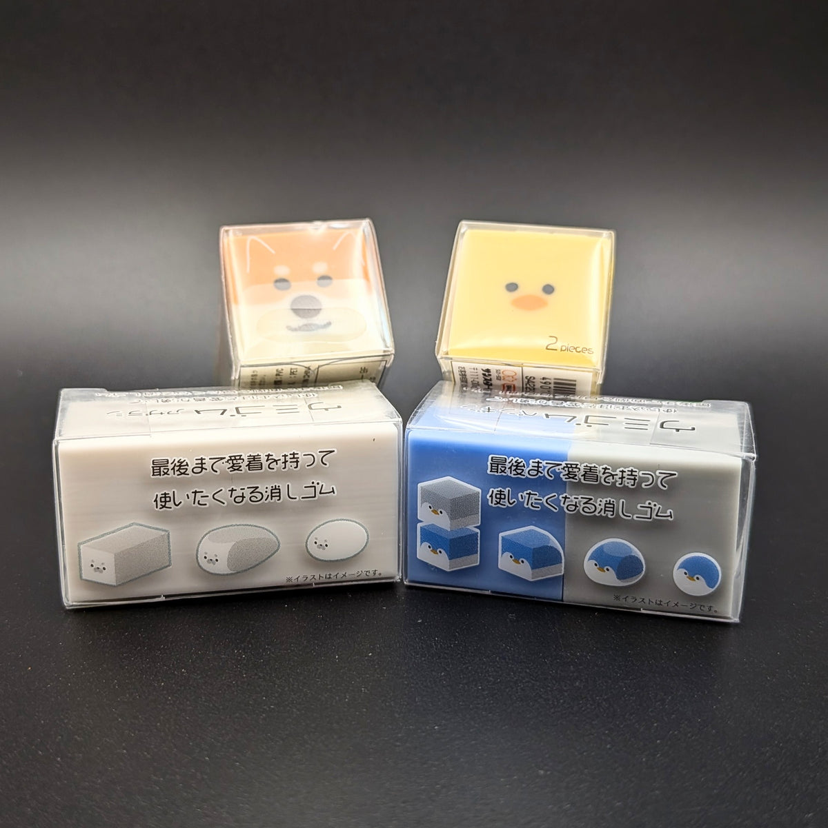 Four small square character-shaped erasers in clear plastic packaging, with Japanese text on the tops and sides. Arranged with two erasers sitting longways, side by side. The other two erasers are leaning up on the others, revealing the faces of a yellow duck and an orange Shiba Inu dog peaking over the top.