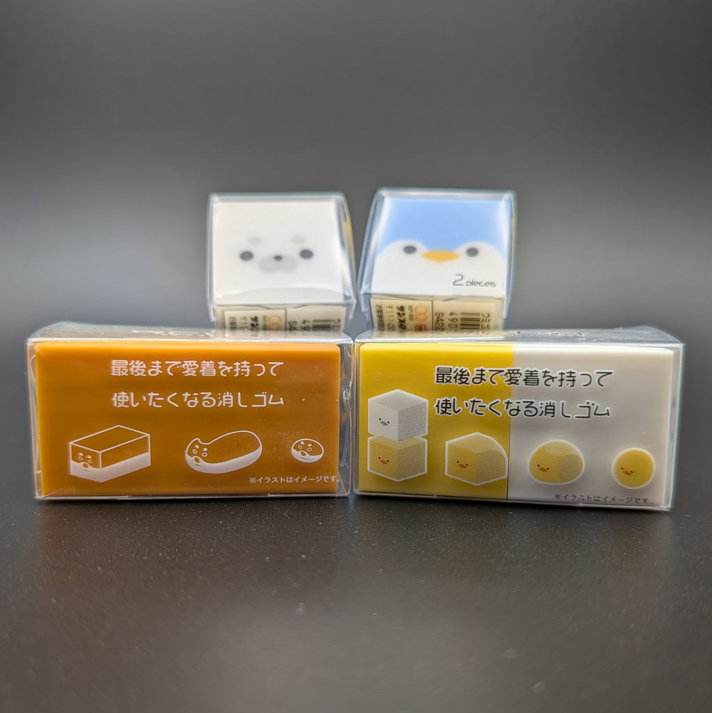 Four small square character-shaped erasers in clear plastic packaging, with Japanese text on the tops and sides. Arranged with two erasers sitting longways, side by side. The other two erasers are leaning up on the others, revealing the faces of a white seal and a blue penguin peaking over the top.