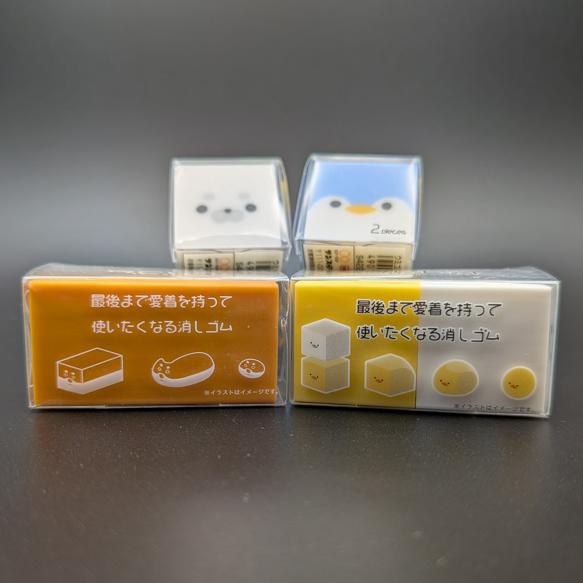 Four small square character-shaped erasers in clear plastic packaging, with Japanese text on the tops and sides. Arranged with two erasers sitting longways, side by side. The other two erasers are leaning up on the others, revealing the faces of a white seal and a blue penguin peaking over the top.