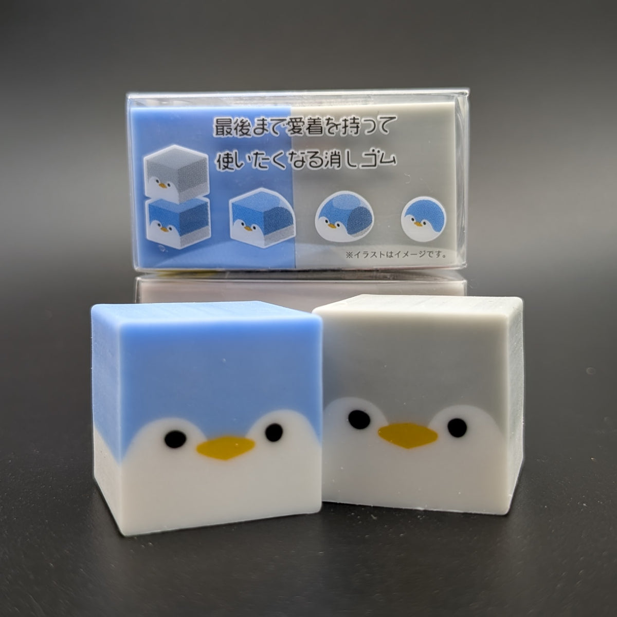 Two small square character-shaped erasers sitting next to each other, one blue penguin and one grey. The side of the package can be seen in the background, showing an image guide of how the eraser works and changes shape over time.