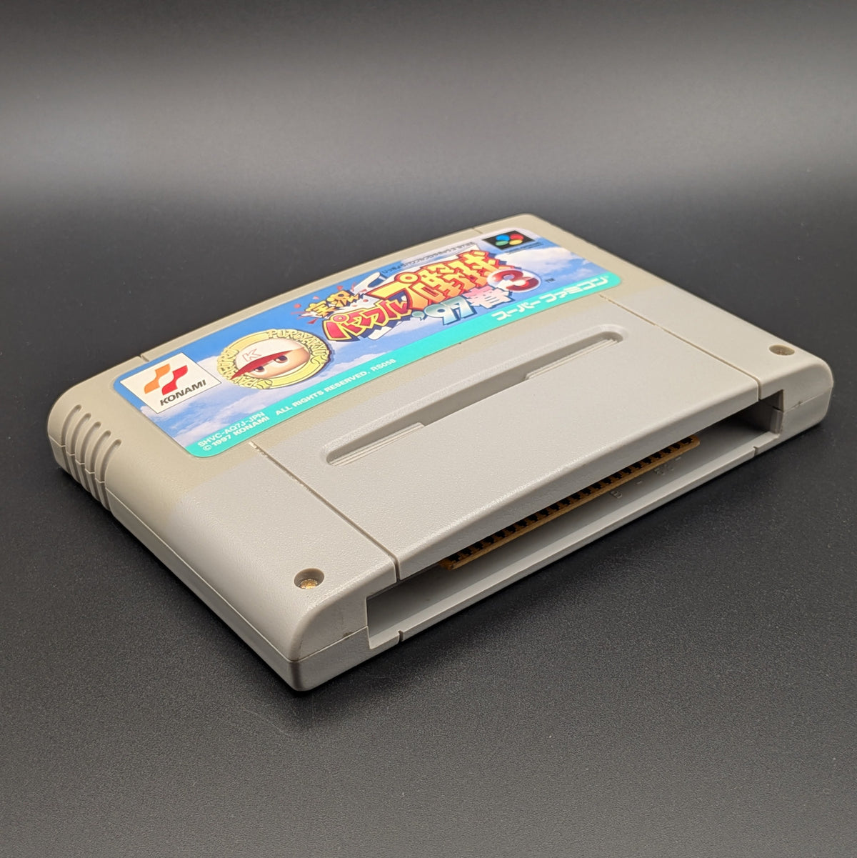 Jikkyou Powerful Pro Yakyuu 3 '97 video game cartridge for Nintendo's Super Famicom (SFC) Entertainment System, shown laying on its back with the label facing upwards. Showing the left hand side and bottom slot. It sits on a dark grey background