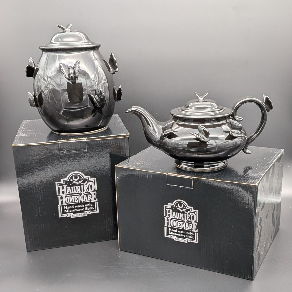 A black ceramic haunted homeware teapot & jar displayed on top of their original Killstar black packaging boxes. both with matching ceramic sculpted butterfly accents on its lid and body.