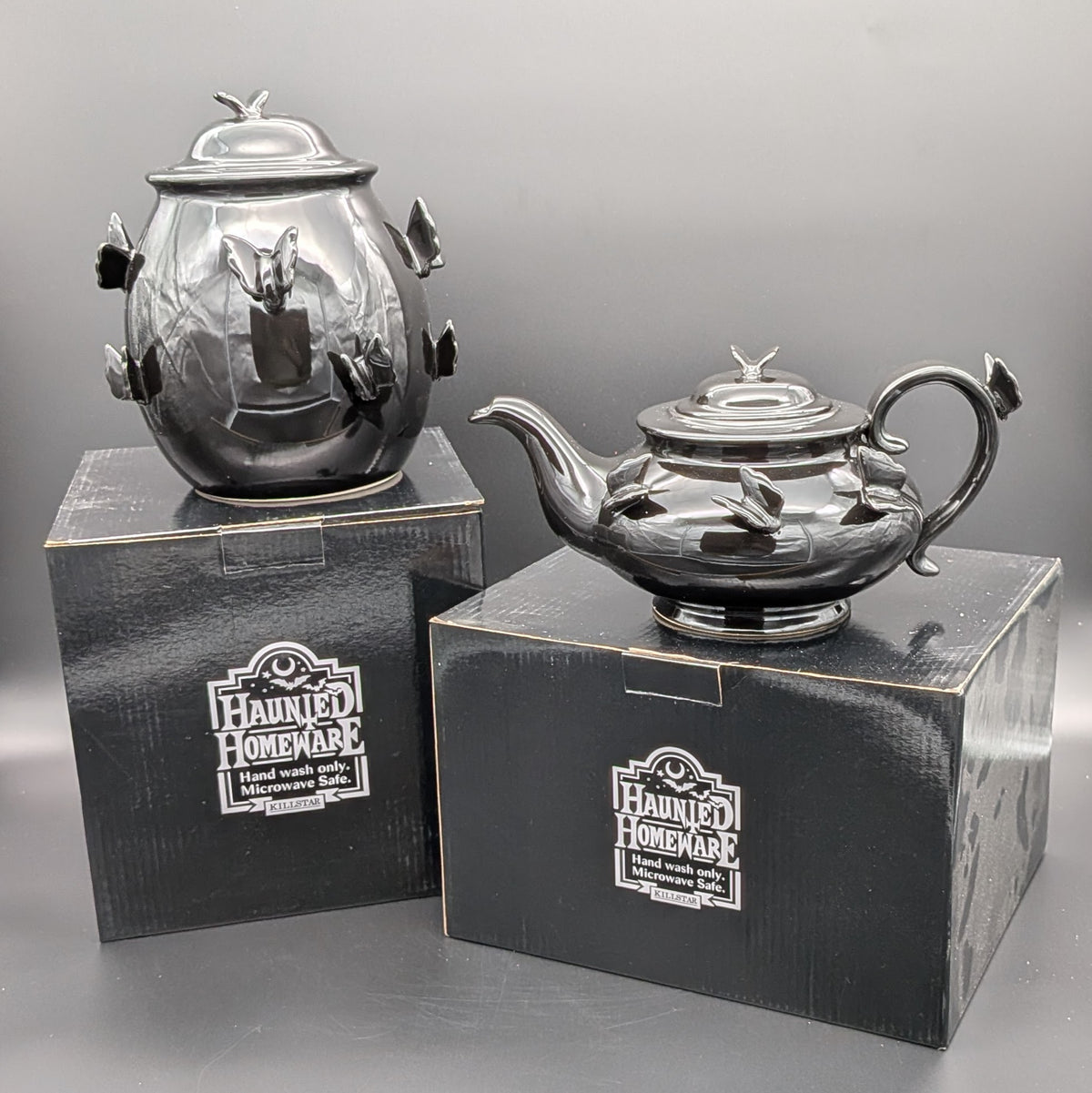 A black ceramic haunted homeware teapot & jar displayed on top of their original Killstar black packaging boxes. both with matching ceramic sculpted butterfly accents on its lid and body.