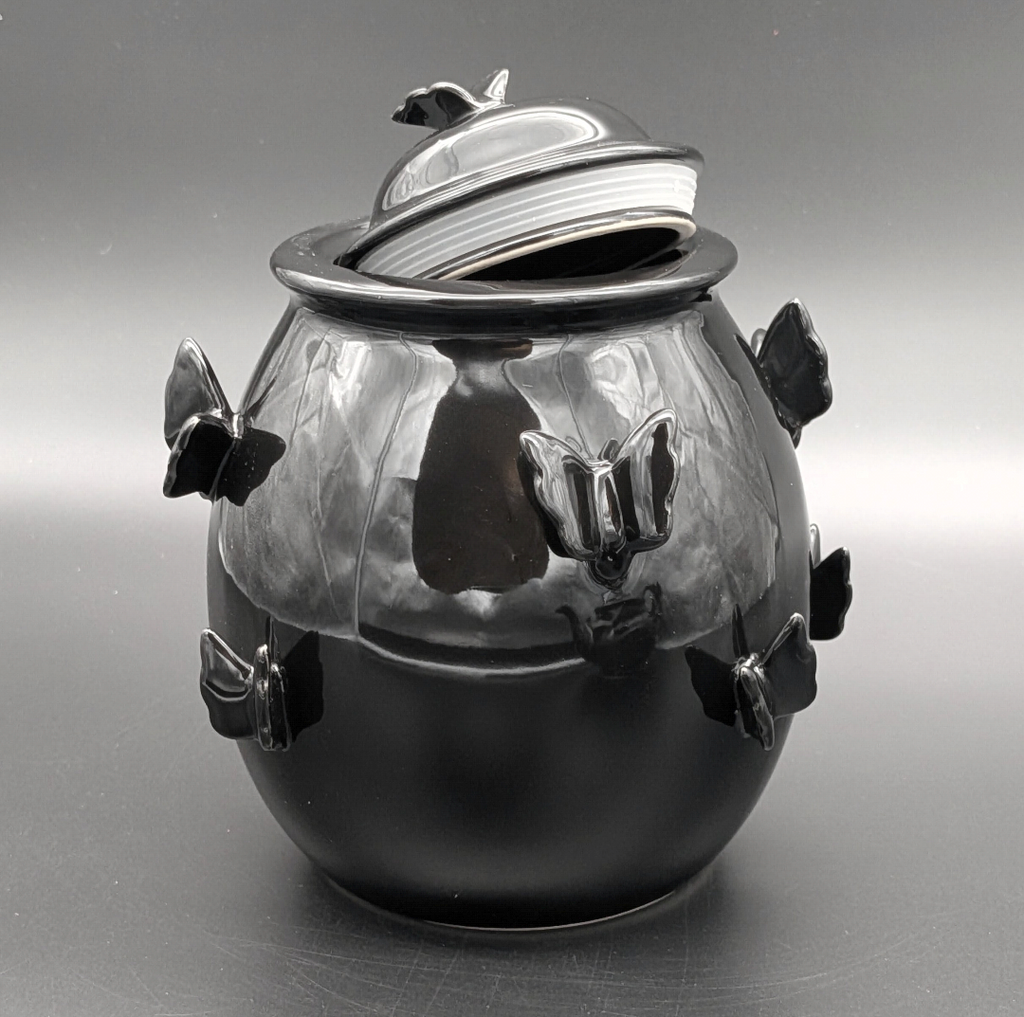 A glossy black ceramic pot with a matching lid, decorated with sculpted butterfly motifs on the sides and on top of the lid, photographed against a neutral grey background.