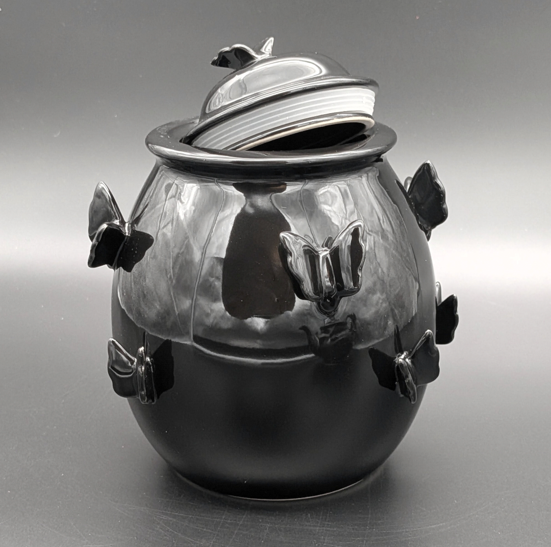 A glossy black ceramic pot with a matching lid, decorated with sculpted butterfly motifs on the sides and on top of the lid, photographed against a neutral grey background.